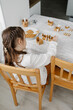 © laniko - Adorable brown-haired girl having hair in two plaits wearing white blose decorate with icing gingerbread houses at the kitchen. Christmas sweets. Gingerbread house - classic holiday tradition