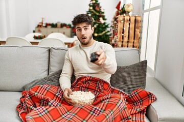 Sticker - Young hispanic man eating popcorn sitting on sofa by christmas tree scared and amazed with open mouth for surprise, disbelief face