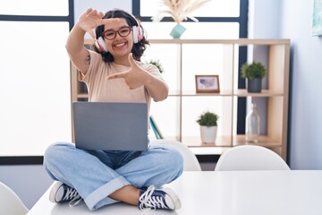 Poster - Young hispanic woman using laptop sitting on the table wearing headphones smiling making frame with hands and fingers with happy face. creativity and photography concept.