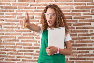 Sticker - Young caucasian woman holding art notebook pointing with finger surprised ahead, open mouth amazed expression, something on the front