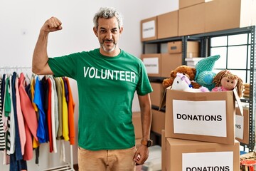 Middle age hispanic man wearing volunteer t shirt at donations stand strong person showing arm muscle, confident and proud of power