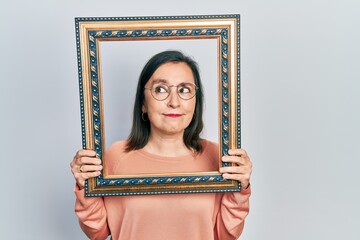 Sticker - Middle age hispanic woman holding empty frame smiling looking to the side and staring away thinking.