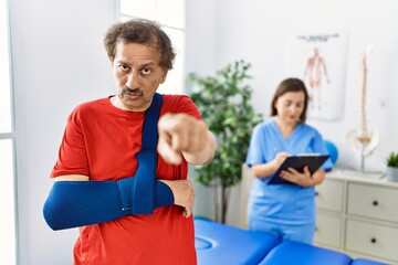 Sticker - Southeast asian man wearing wearing arm on sling at rehabilitation clinic pointing with finger to the camera and to you, confident gesture looking serious