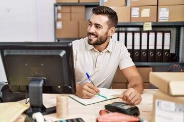 Young hispanic man smiling confident writing on notebook at storehouse