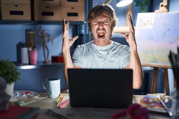 Sticker - Middle age man sitting at art studio with laptop at night crazy and mad shouting and yelling with aggressive expression and arms raised. frustration concept.