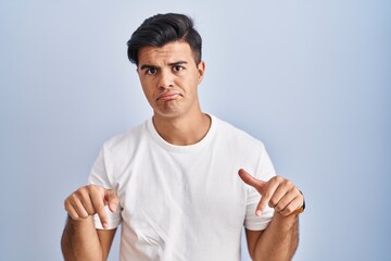 Sticker - Hispanic man standing over blue background pointing down looking sad and upset, indicating direction with fingers, unhappy and depressed.