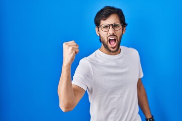 Poster - Handsome latin man standing over blue background angry and mad raising fist frustrated and furious while shouting with anger. rage and aggressive concept.