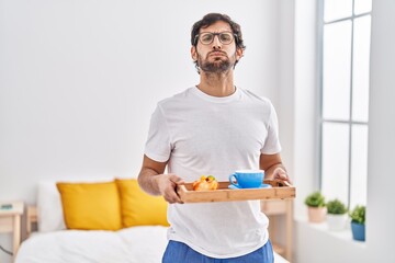 Wall Mural - Handsome latin man eating breakfast on the bed puffing cheeks with funny face. mouth inflated with air, catching air.