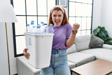 Wall Mural - Young redhead woman holding recycling wastebasket with plastic bottles very happy and excited doing winner gesture with arms raised, smiling and screaming for success. celebration concept.