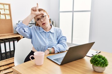 Poster - Young redhead woman working at the office using computer laptop making fun of people with fingers on forehead doing loser gesture mocking and insulting.