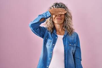 Wall Mural - Middle age woman standing over pink background smiling and laughing with hand on face covering eyes for surprise. blind concept.