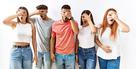 Wall Mural - Group of young friends standing together over isolated background peeking in shock covering face and eyes with hand, looking through fingers with embarrassed expression.