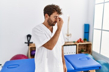 Canvas Print - Young handsome physiotherapist man working at pain recovery clinic tired rubbing nose and eyes feeling fatigue and headache. stress and frustration concept.