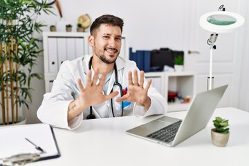 Wall Mural - Young doctor working at the clinic using computer laptop afraid and terrified with fear expression stop gesture with hands, shouting in shock. panic concept.