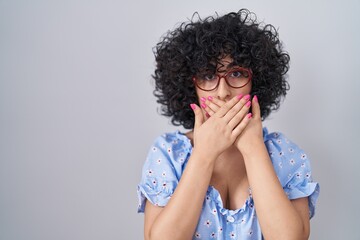 Canvas Print - Young brunette woman with curly hair wearing glasses over isolated background shocked covering mouth with hands for mistake. secret concept.