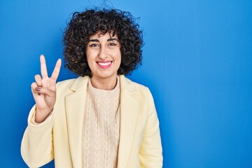 Sticker - Young brunette woman with curly hair standing over blue background showing and pointing up with fingers number two while smiling confident and happy.