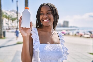 Sticker - Young african american girl smiling happy holding sunscreen lotion at the promenade.