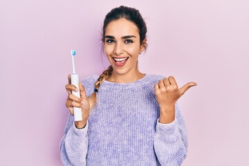 Wall Mural - Young hispanic girl holding electric toothbrush pointing thumb up to the side smiling happy with open mouth