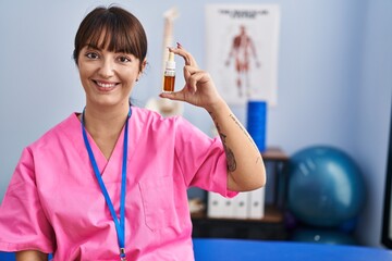 Poster - Young brunette woman holding cbd oil at physiotherapy clinic looking positive and happy standing and smiling with a confident smile showing teeth