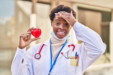 Wall Mural - African american woman wearing doctor uniform holding heart stressed and frustrated with hand on head, surprised and angry face