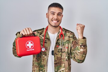 Poster - Young hispanic doctor wearing camouflage army uniform holding first aid kit screaming proud, celebrating victory and success very excited with raised arms