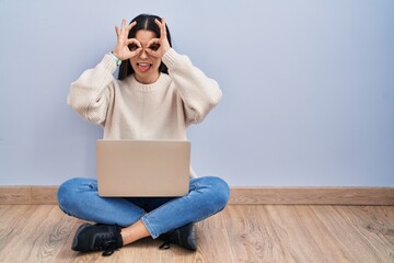 Poster - Young woman using laptop sitting on the floor at home doing ok gesture like binoculars sticking tongue out, eyes looking through fingers. crazy expression.