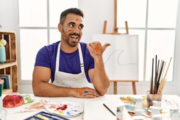 Poster - Young hispanic man with beard at art studio with painted face smiling with happy face looking and pointing to the side with thumb up.