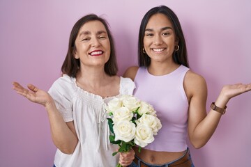 Poster - Hispanic mother and daughter holding bouquet of white flowers smiling cheerful presenting and pointing with palm of hand looking at the camera.