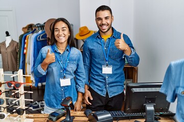 Wall Mural - Young interracial people working at retail boutique doing happy thumbs up gesture with hand. approving expression looking at the camera showing success.