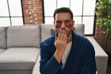 young hispanic man with beard wearing beauty eyes patch covering mouth with hand, shocked and afraid