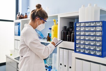 Poster - Young blonde woman wearing scientist uniform and medical mask writing on clipboard at laboratory