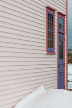 The Exterior Wall Of A White Wooden Cape Cod Clapboard Siding House With A Purple Panel Door And Vintage Glass Window, Black Metal Hinges, Pink Trim Around The Door, And White Snow On The Step. 