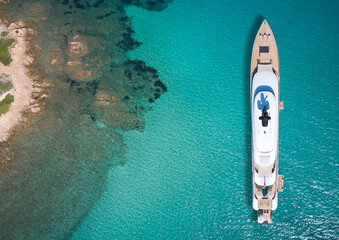 Wall Mural - White big yacht for a billionaire anchored off a rocky shore aerial view. Mega yacht on blue water aerial view. Modern big white yacht on transparent water, coastline top view.