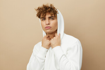 a stylish young man in a white hoodie, standing on a beige background and holding the hood on his head with his hands, smiling pleasantly at the camera