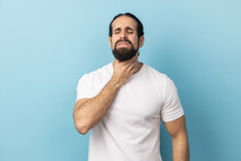 Portrait Of Unhealthy Man With Beard Wearing White T-shirt Suffering From Sore Throat, Keeping Hand On Her Neck, Frowning Face And Closed Eyes. Indoor Studio Shot Isolated On Blue Background.