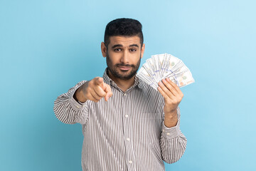 Portrait of young businessman pointing to camera and holding dollar banknotes, encouraging to win lottery, earn profit, wearing striped shirt. Indoor studio shot isolated on blue background.