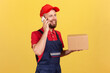 © khosrork - Portrait of positive courier woman standing holding cardboard box, parcel for client and talking via cell phone, wearing overalls and red cap. Indoor studio shot isolated on yellow background.