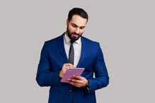 Portrait Of Positive Bearded Businessman Writing Down In Paper Notebook, Making To Do List, Having Good Mood, Wearing Official Style Suit. Indoor Studio Shot Isolated On Gray Background.