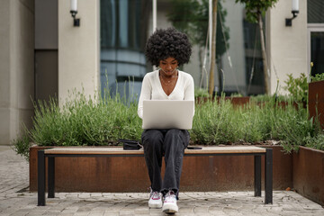 Wall Mural - Young african american businesswoman working using laptop sitting on the bench in the city