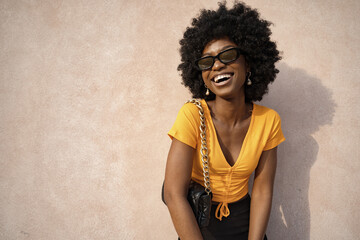 Wall Mural - Happy African American young woman with afro hair wearing stylish sunglasses posing against beige wall background