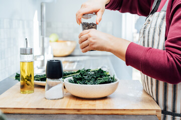 Close up woman adding spices and herbs while cooking kale chips or healthy salad for dinner on the kitchen table. Healthy eating, dieting lifestyle. Selective focus, copy space.