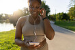 © gpointstudio - African American woman using mobile phone and earphones before exercising at the park in a  summer day