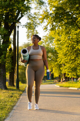 Wall Mural - Plus sized African American woman walking with exercise mat and water through the park in a summer day