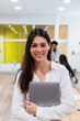 © Westend61 - Portrait of smiling businesswoman holding laptop in office with colleagues in background
