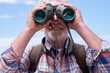 © luciano - Close up on active senior man  in outdoors excursion using binoculars looking at camera, elderly caucasian male enjoying freedom and healthy lifestyle