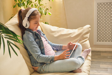 Wall Mural - Cute little girl in casual clothes and headphones using a tablet