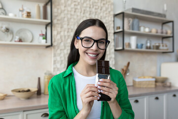 Young beautiful woman in glasses and a green shirt at home in the kitchen, holding chocolate in her hands, eating a bar of black chocolate. Look at the camera, smile.