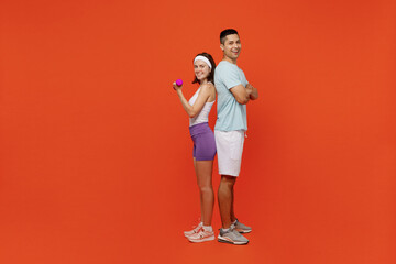 Full body side view young fitness instructor sporty two man woman in headband t-shirt stand back to back hold dumbbell spend weekend in gym isolated on plain orange background. Workout sport concept.