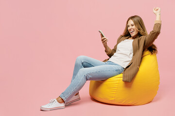 Full body young fun successful employee business woman 30s she wear casual classic jacket sit in bag chair hold use mobile cell phone do winner gesture isolated on plain pastel light pink background.