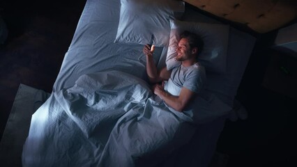 Leinwandbilder - Top View Apartment Bedroom: Handsome Young Man Using Smartphone in Bed at Night. Smiling Guy Browsing Social Media, Dating Apps, Doing Internet Online Shopping on Mobile Phone. Top Down Zoom Out